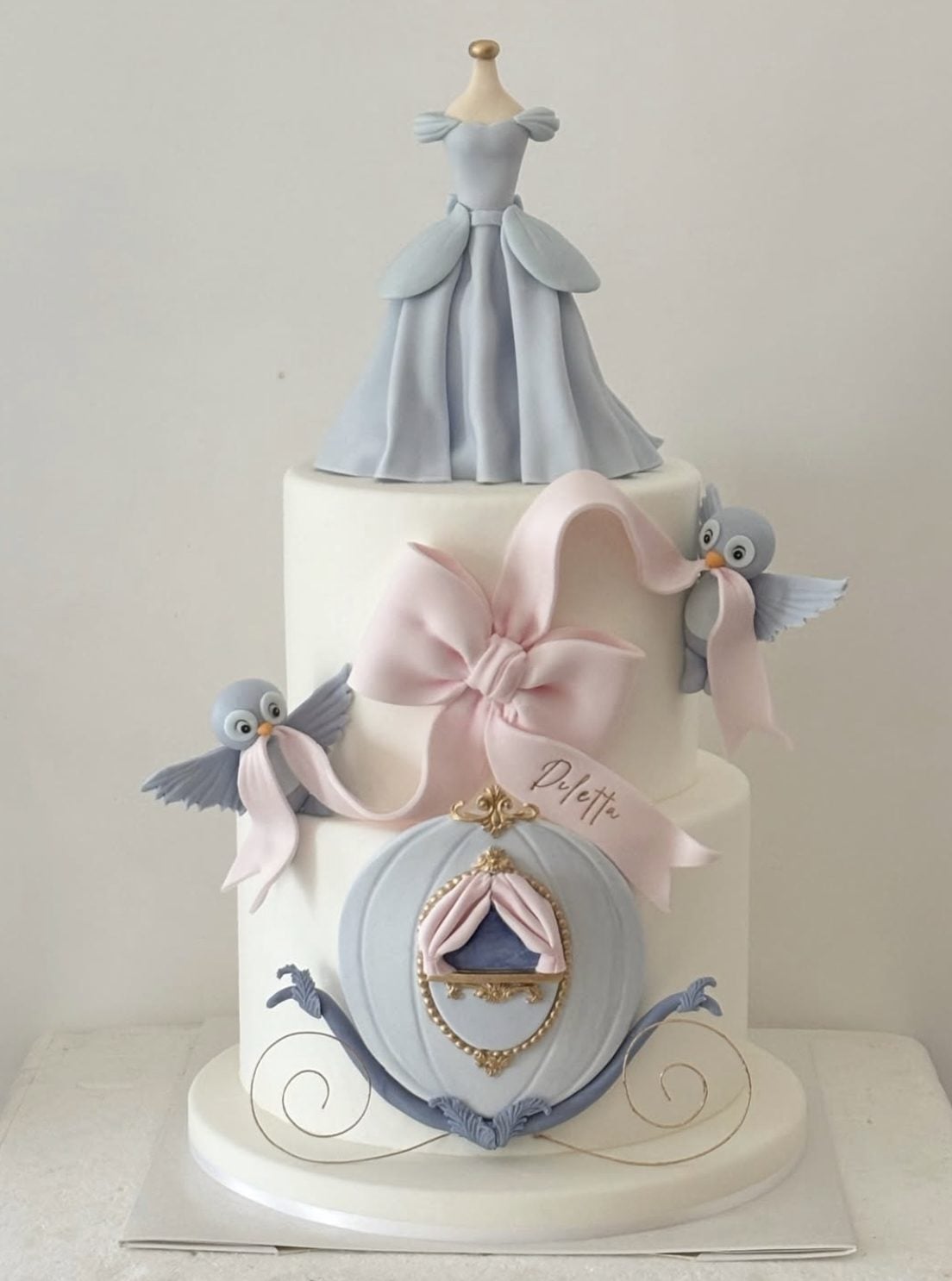 This Cinderella birthday cake is soft, elegant, and full of beautiful fairytale details. The white tiers keep the design clean and classic, while the pale blue dress topper, pumpkin carriage, pink bow, and sweet bird accents make the cake feel completely magical.