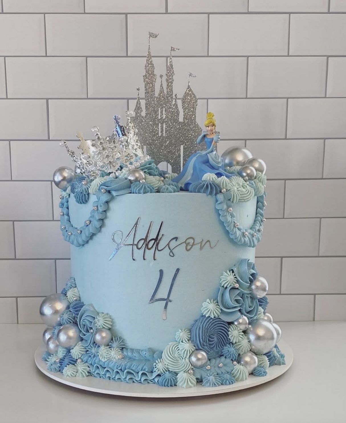 This Cinderella birthday cake is a soft blue buttercream with a silver castle topper, Cinderella figurine, pearl details, and oversized silver accents.