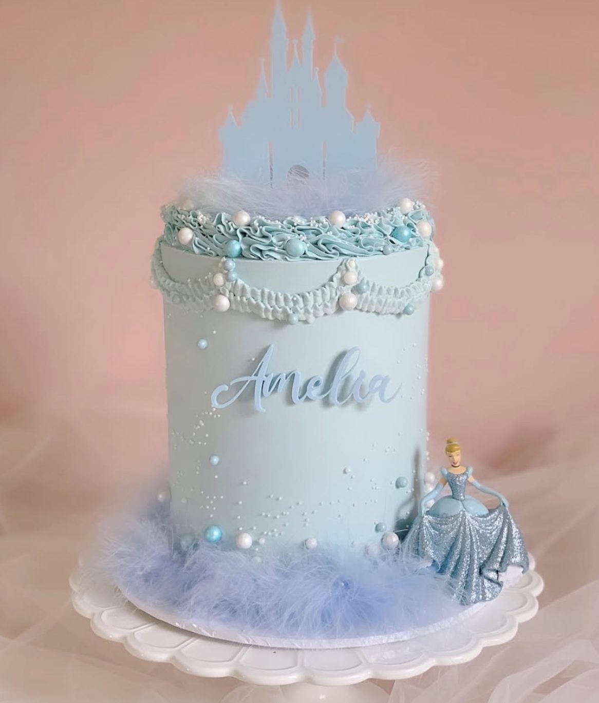 This Cinderella birthday cake is soft, sparkly, and so elegant. The pale blue frosting, white castle topper, pearl accents, delicate piped details, fluffy trim, and Cinderella figurine give it a dreamy, royal ball look without making the cake feel overdone.