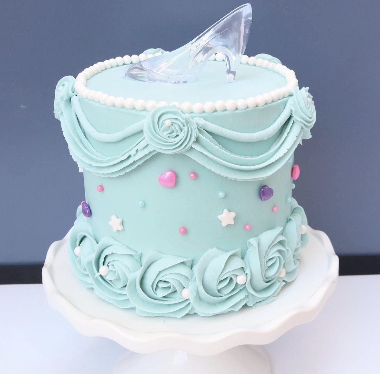 This Cinderella glass slipper cake is simple, sweet, and instantly recognizable. The pale blue frosting, pearl border, soft buttercream swirls, tiny star and heart details, and clear glass slipper topper make it feel magical without needing a complicated design.