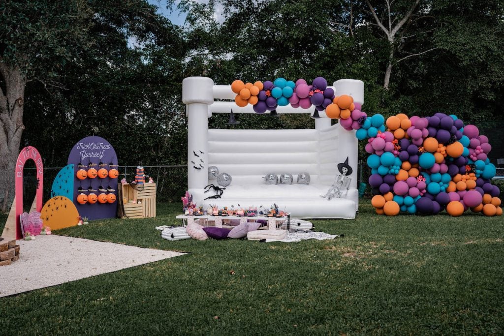 Colorful Halloween Picnic Party with a white bounce house, orange, blue, pink, and purple balloon garland, and other Halloween-inspired decorations